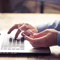 Eine Hand tippt auf dem Laptop, eine zweite Hand hält ein Schloss aus Plastik.